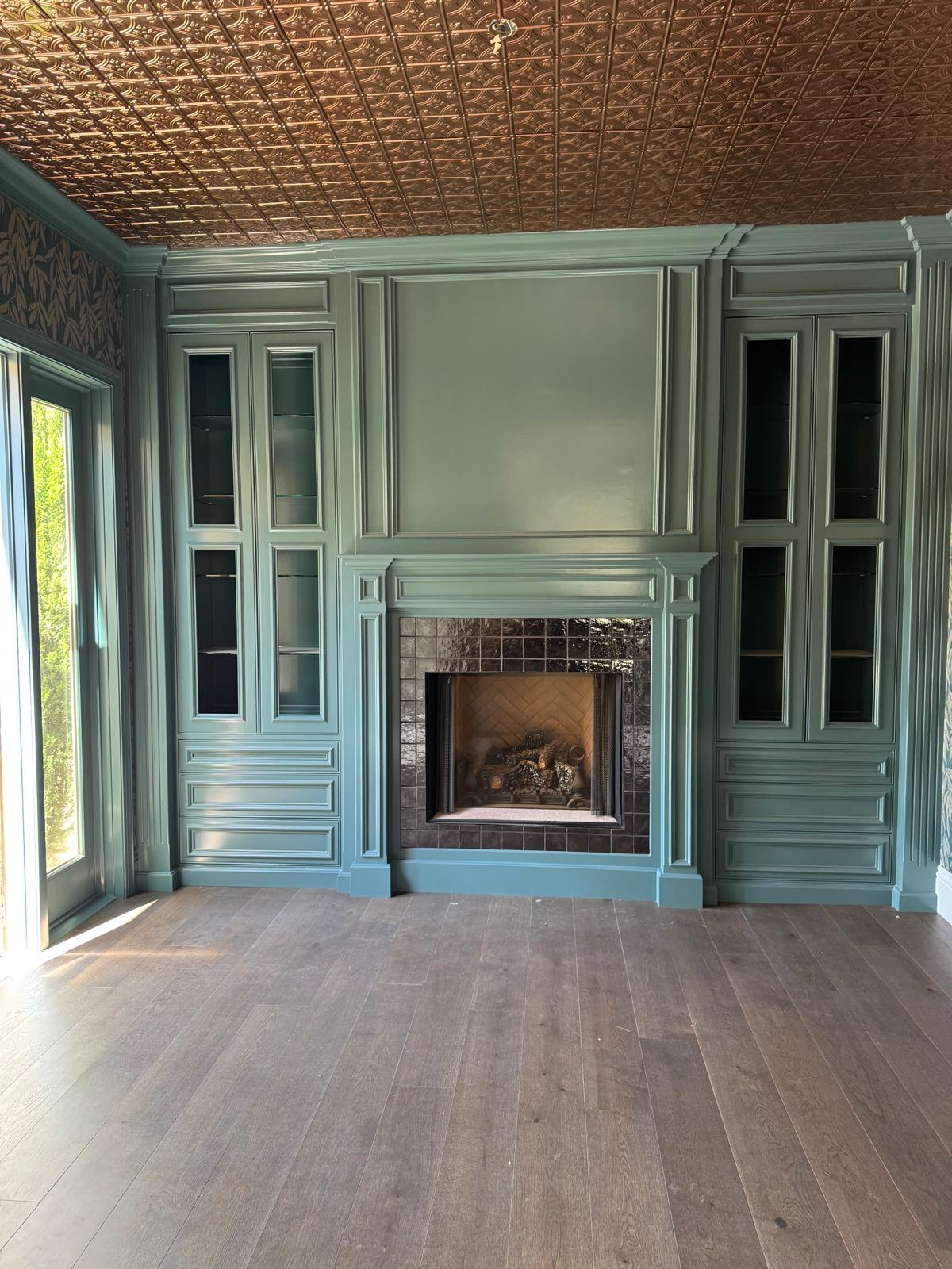 Sage green painted fireplace wall with a moulded mantel, raised-panel surround and matching glass-front built-in cabinets with drawer banks on either side, under a pressed tin ceiling