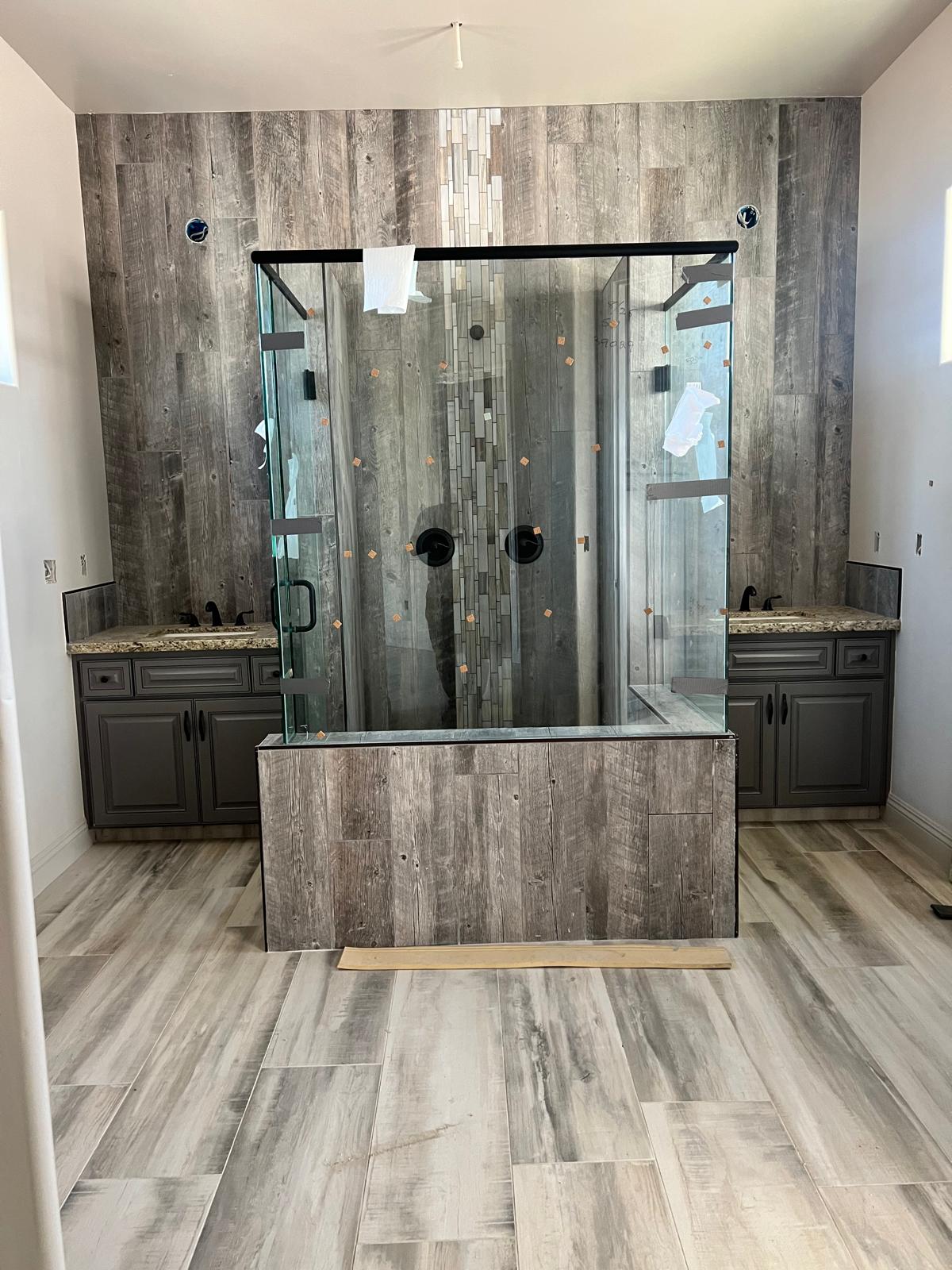 Two charcoal raised-panel vanities with granite tops and matte black faucets flanking a glass walk-in shower framed in barnwood-look tile