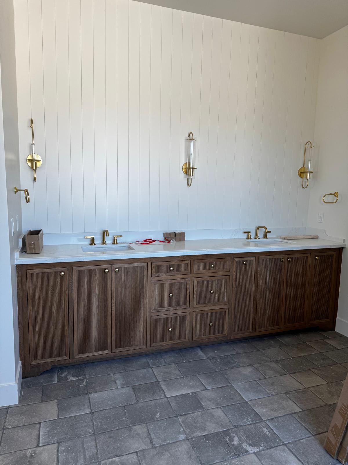 Long double vanity in reeded walnut-toned ash with a white quartz top, brass sconces and shiplap wall