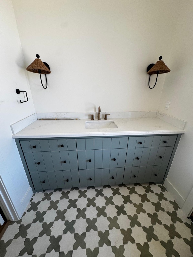 Custom sage green bathroom vanity with vertical-groove drawer banks, matte black knobs, white veined quartz countertop, brass faucet, and woven bell sconces above geometric floor tile