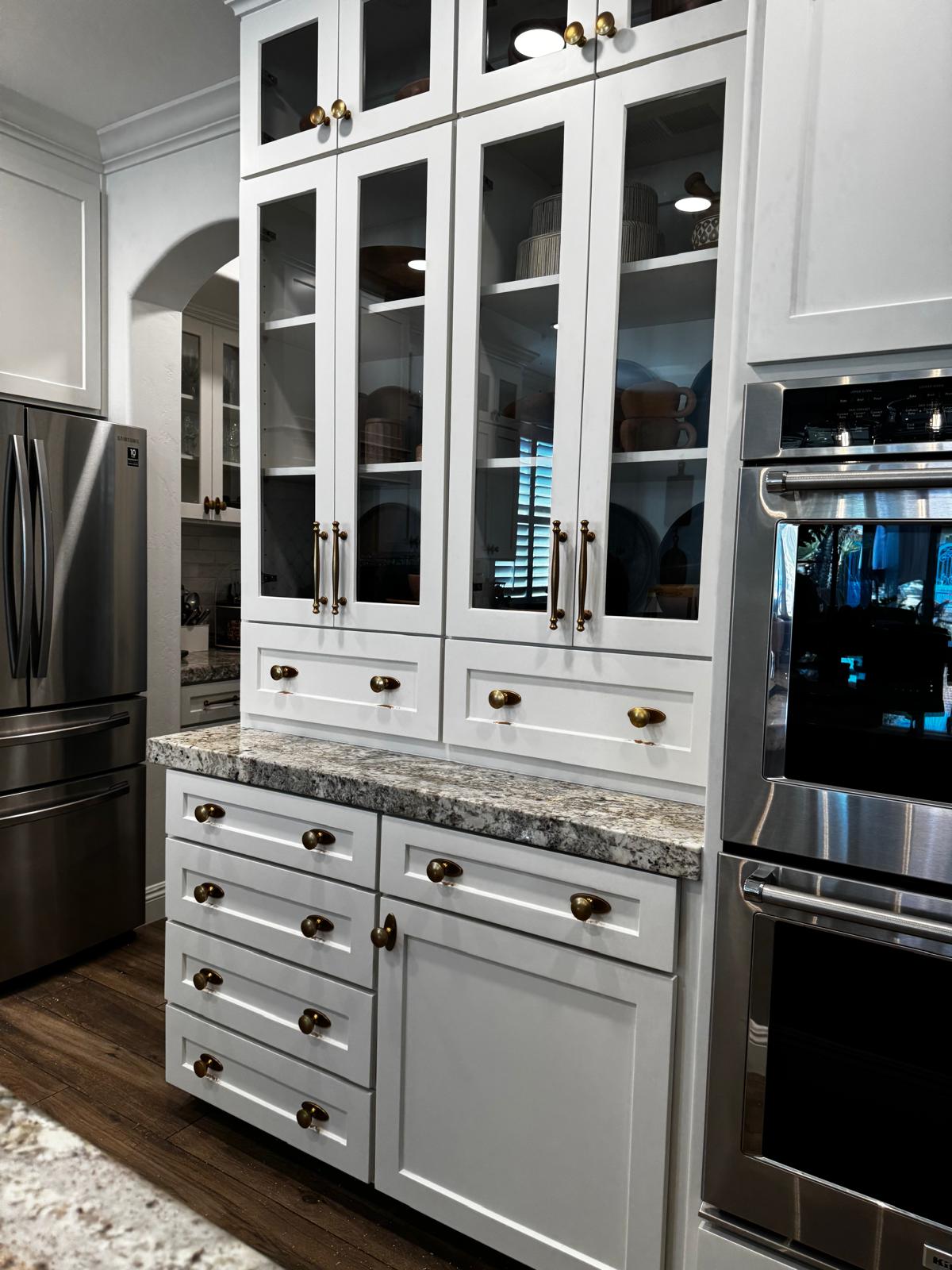 Built-in white shaker china hutch with tall glass-front upper doors, antique brass cup pulls and knobs, a granite counter and a bank of drawers below
