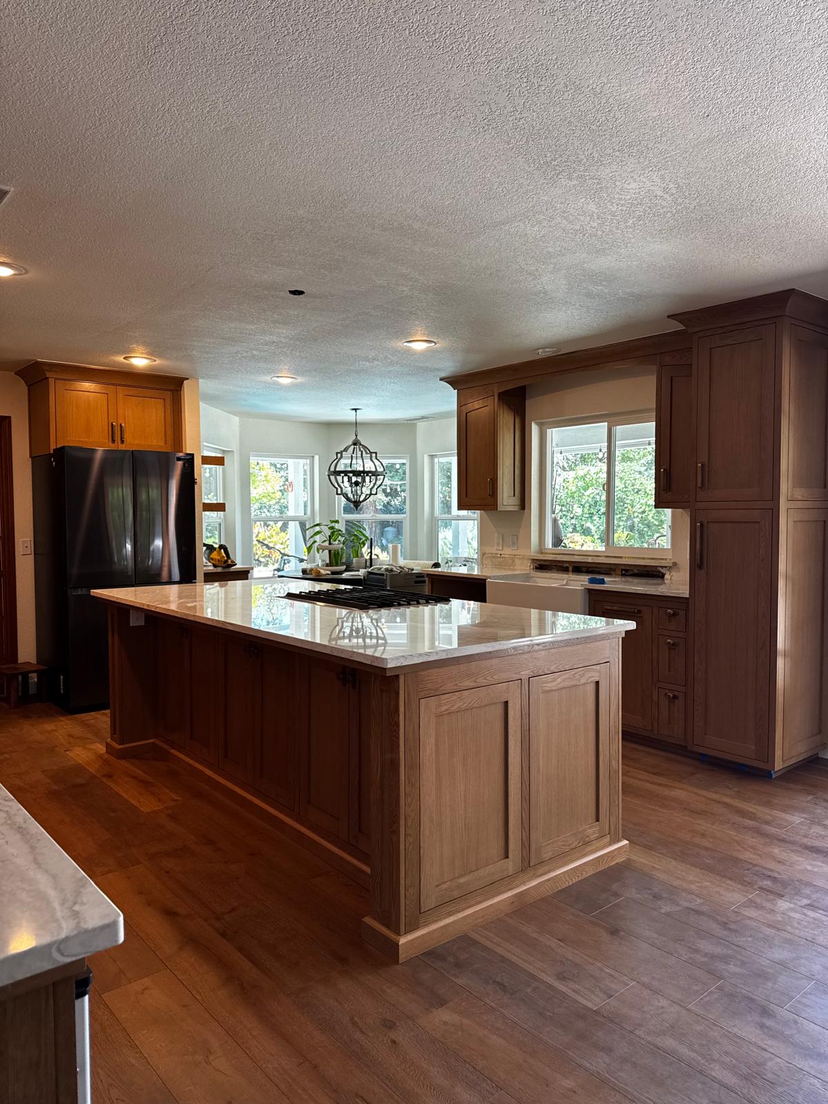 Natural white oak shaker kitchen with a long island under a marble-look quartz top, a gas cooktop, farmhouse sink and a bay-window dining nook beyond