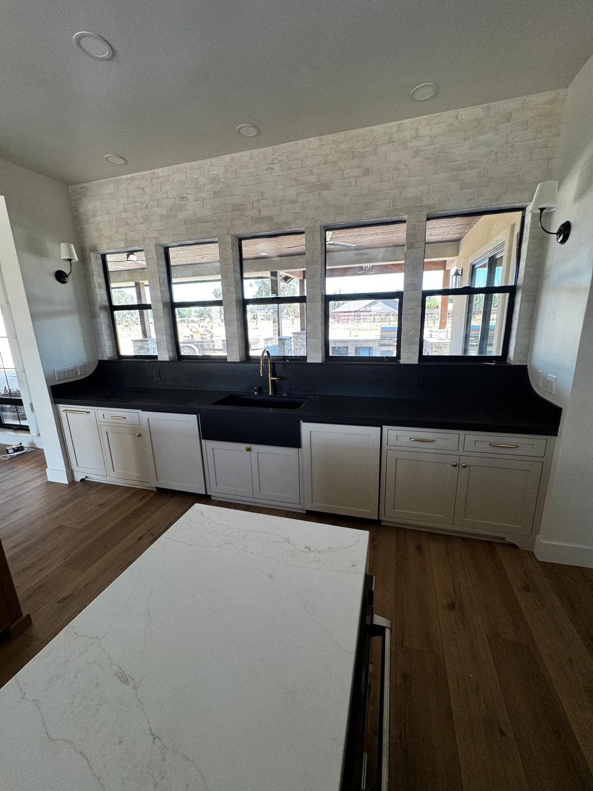 White inset shaker cabinetry running beneath five black-framed windows with a black stone counter, apron sink and brass faucet
