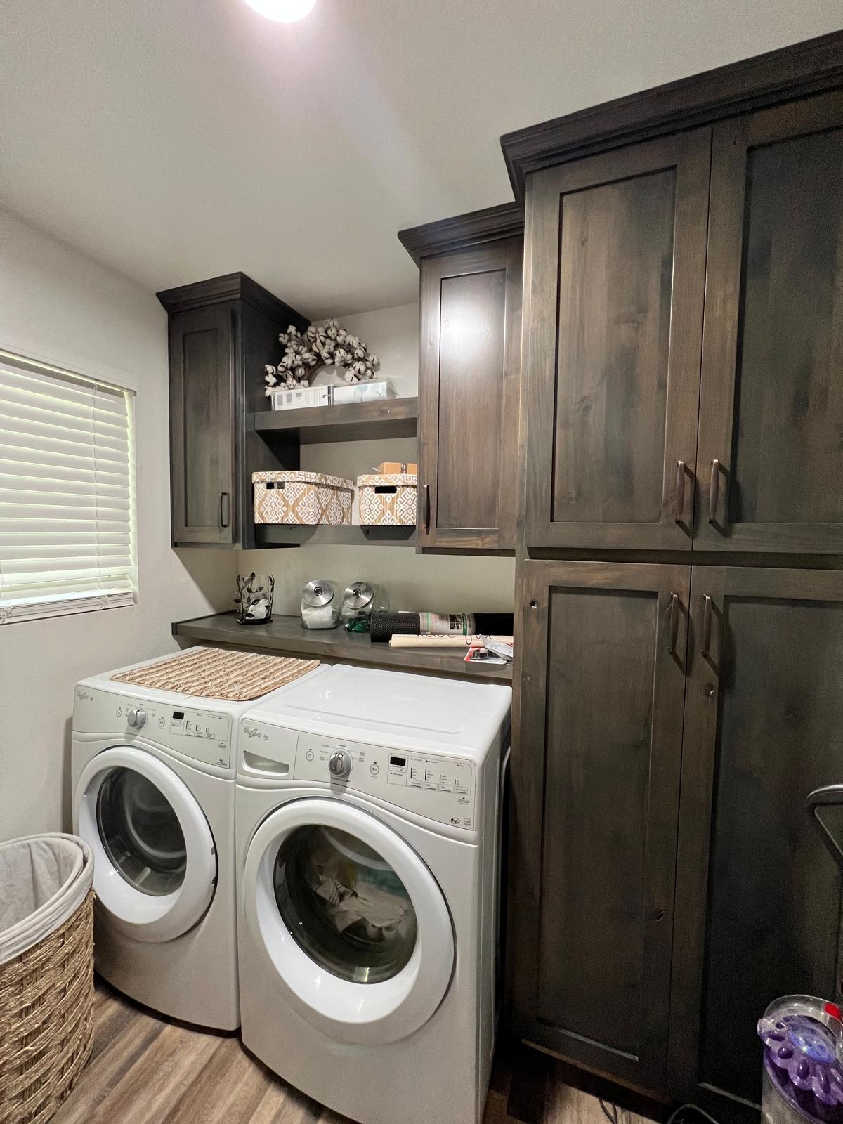 Dark stained alder laundry cabinetry with a floor-to-ceiling storage tower, crown-topped uppers, floating shelves and a folding counter spanning the washer and dryer