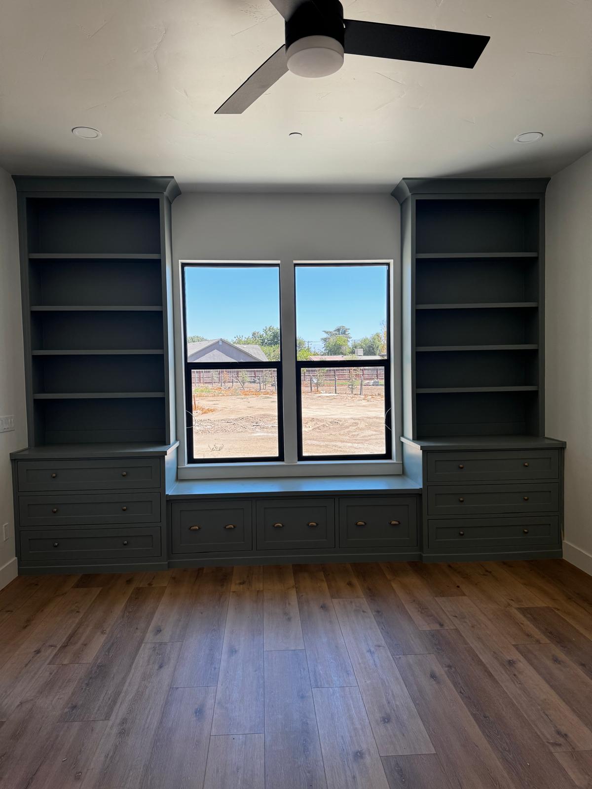 Slate green built-in bookcase wall with open shelving towers, drawer banks and a window seat bench between them
