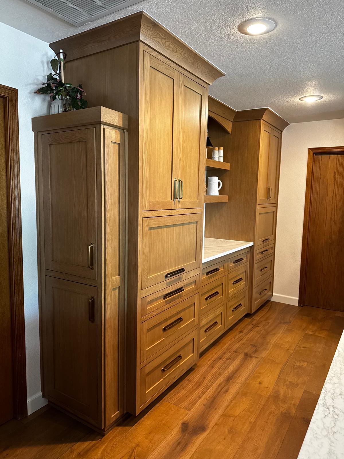 Run of white oak pantry towers with antique brass pulls flanking a coffee bar of stacked drawers, a stone counter and an arched niche with floating shelves