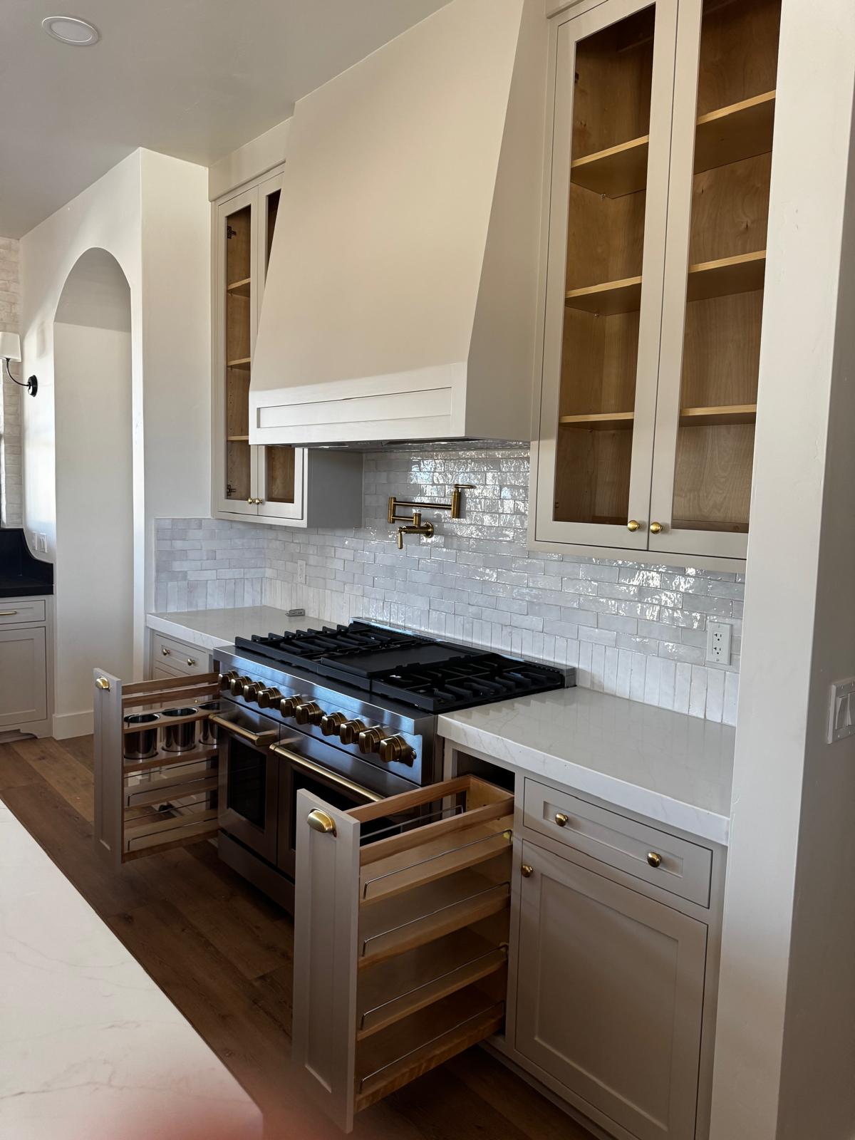 Greige inset cabinetry around a pro range with a custom hood, zellige backsplash, glass-front uppers and two maple pull-out racks extended
